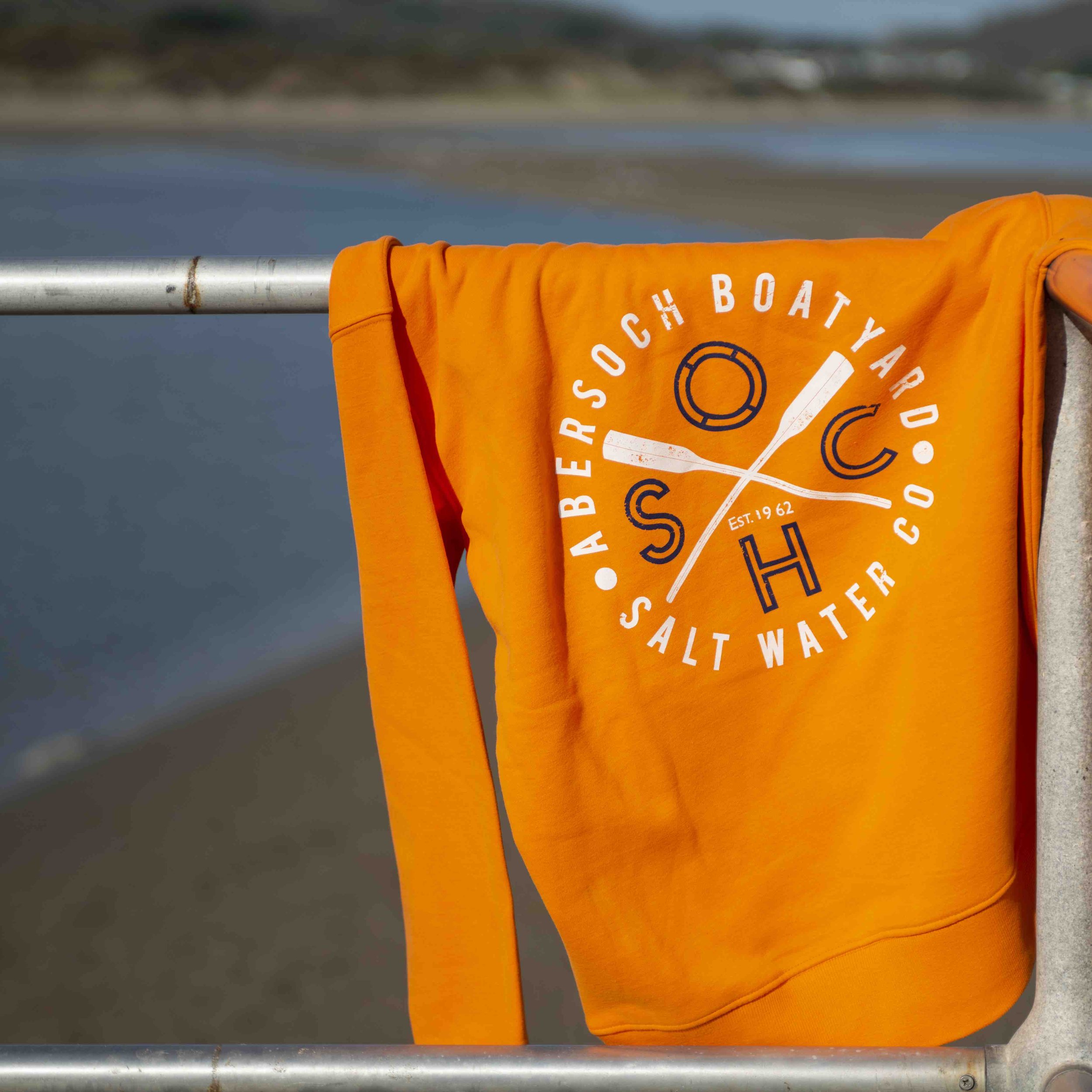 Orange sweatshirt with "ABERSOCH BOATYARD CO SALT WATER" logo on railing near water