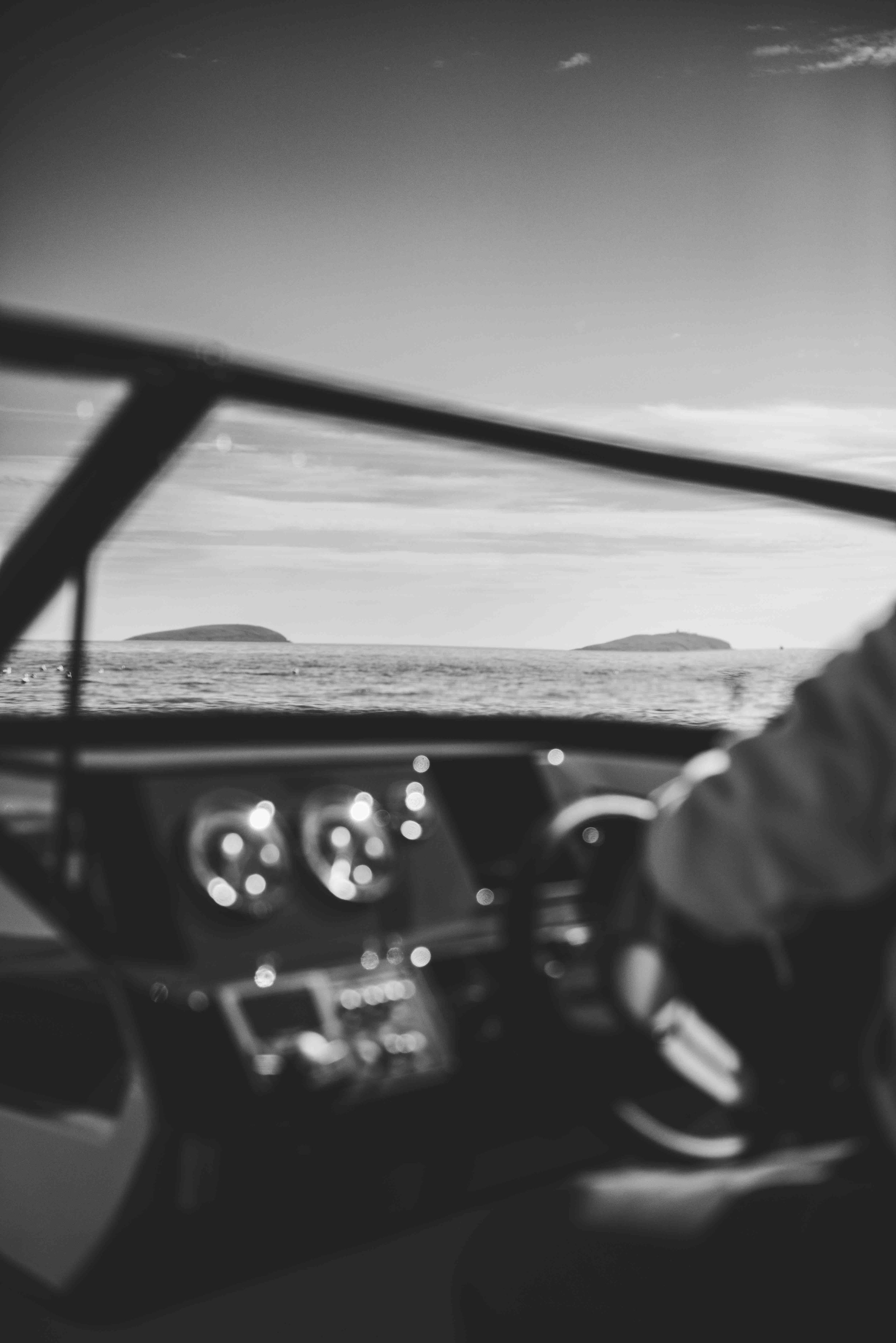 Black and white image of a boat cockpit with a steering wheel and dashboard, overlooking a body of water with distant islands.
