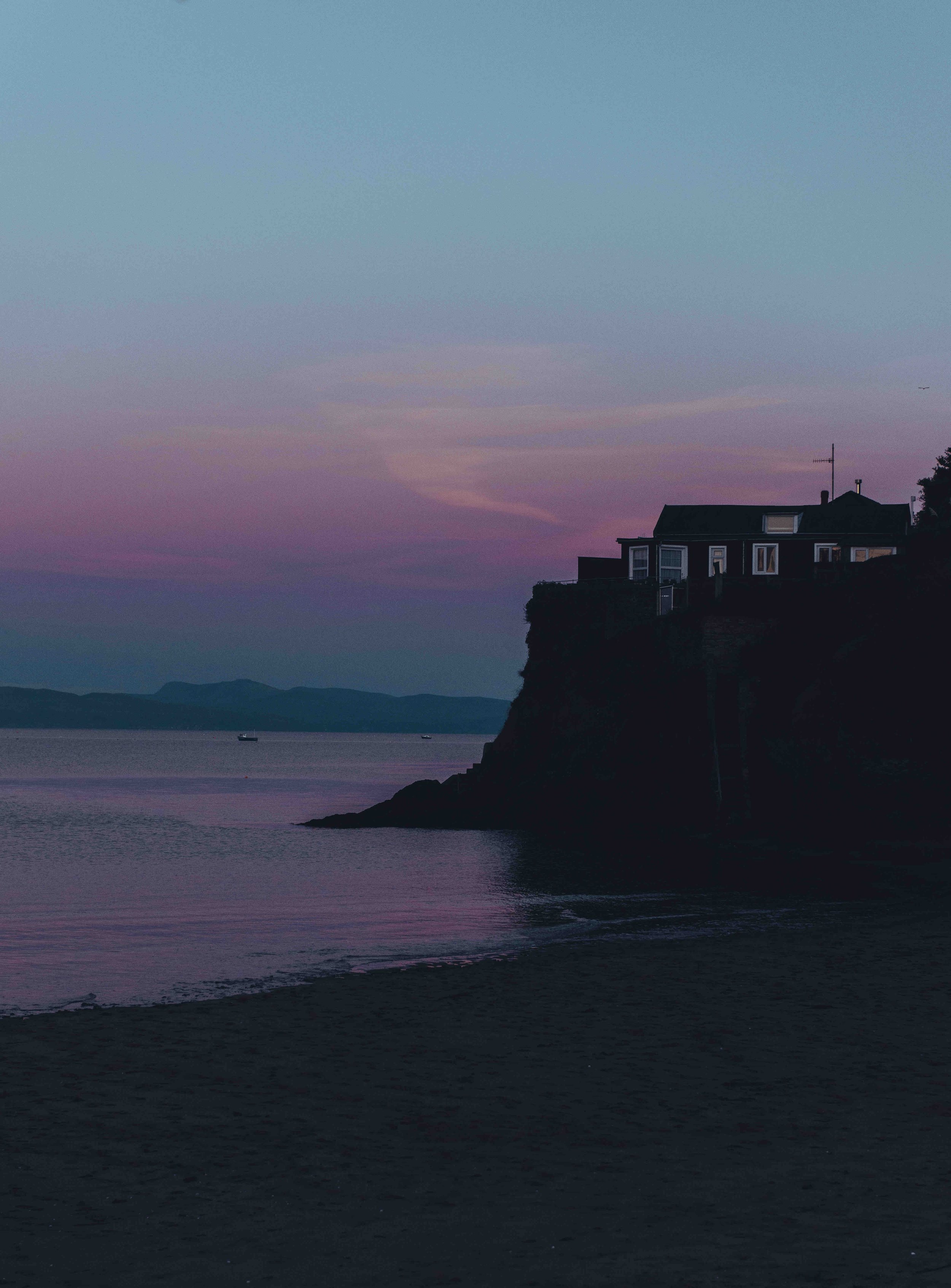 Coastal house silhouette at sunset with colorful sky, ocean, and distant hills.