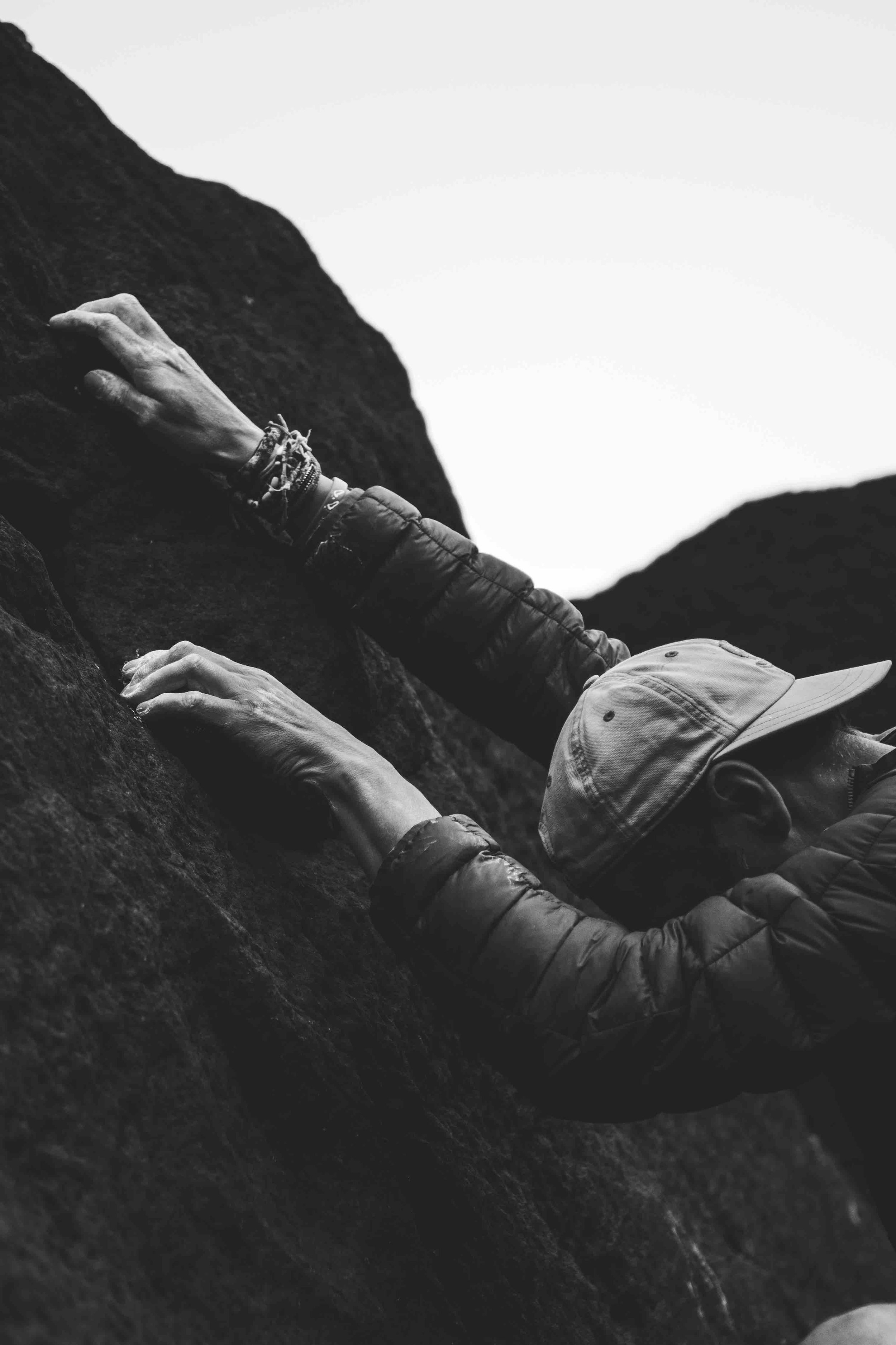 Person rock climbing outdoors, wearing a cap and jacket, hands gripping the rock surface, black and white photo.