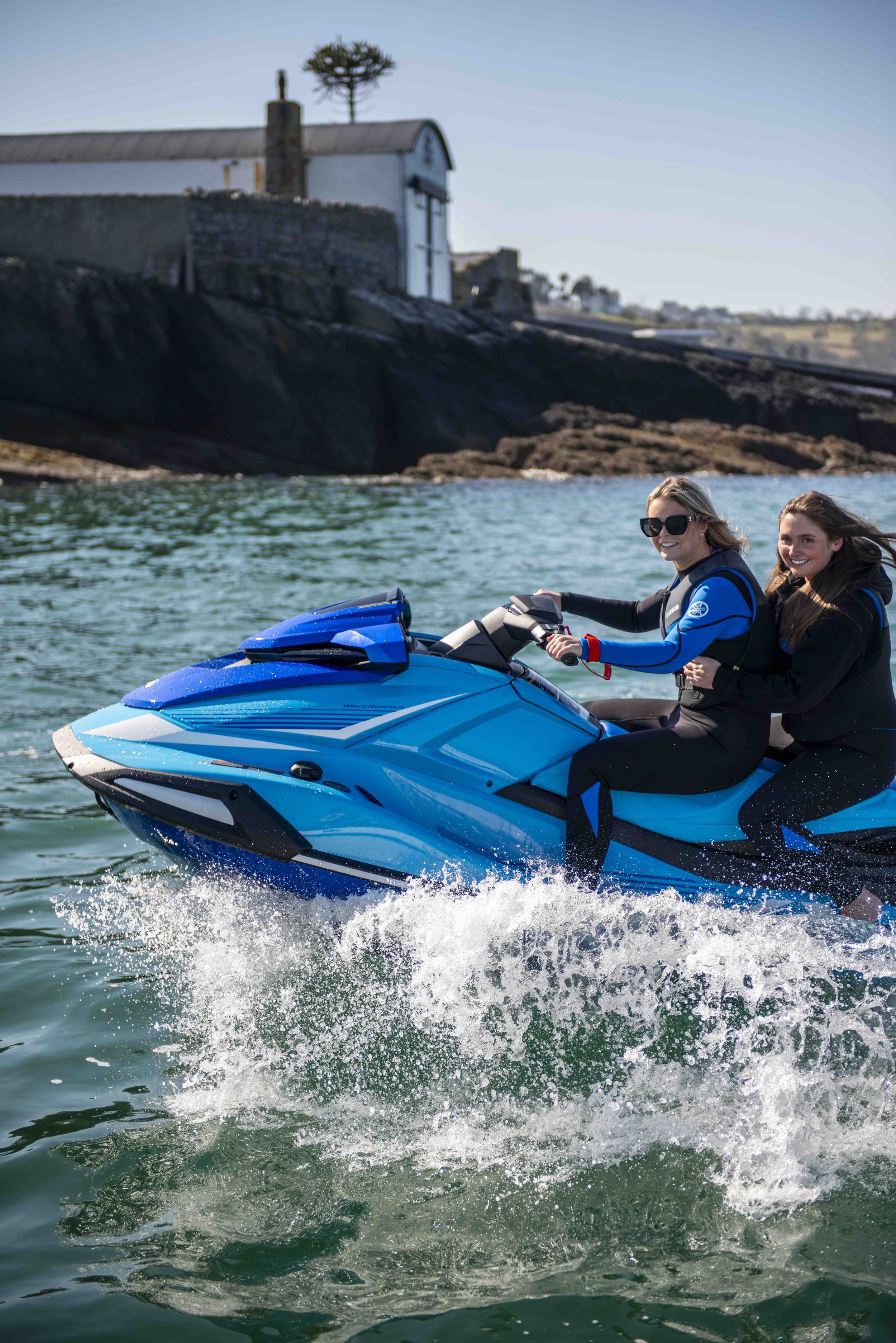 Two people riding a blue jet ski on the water near a rocky shore with a building in the background.