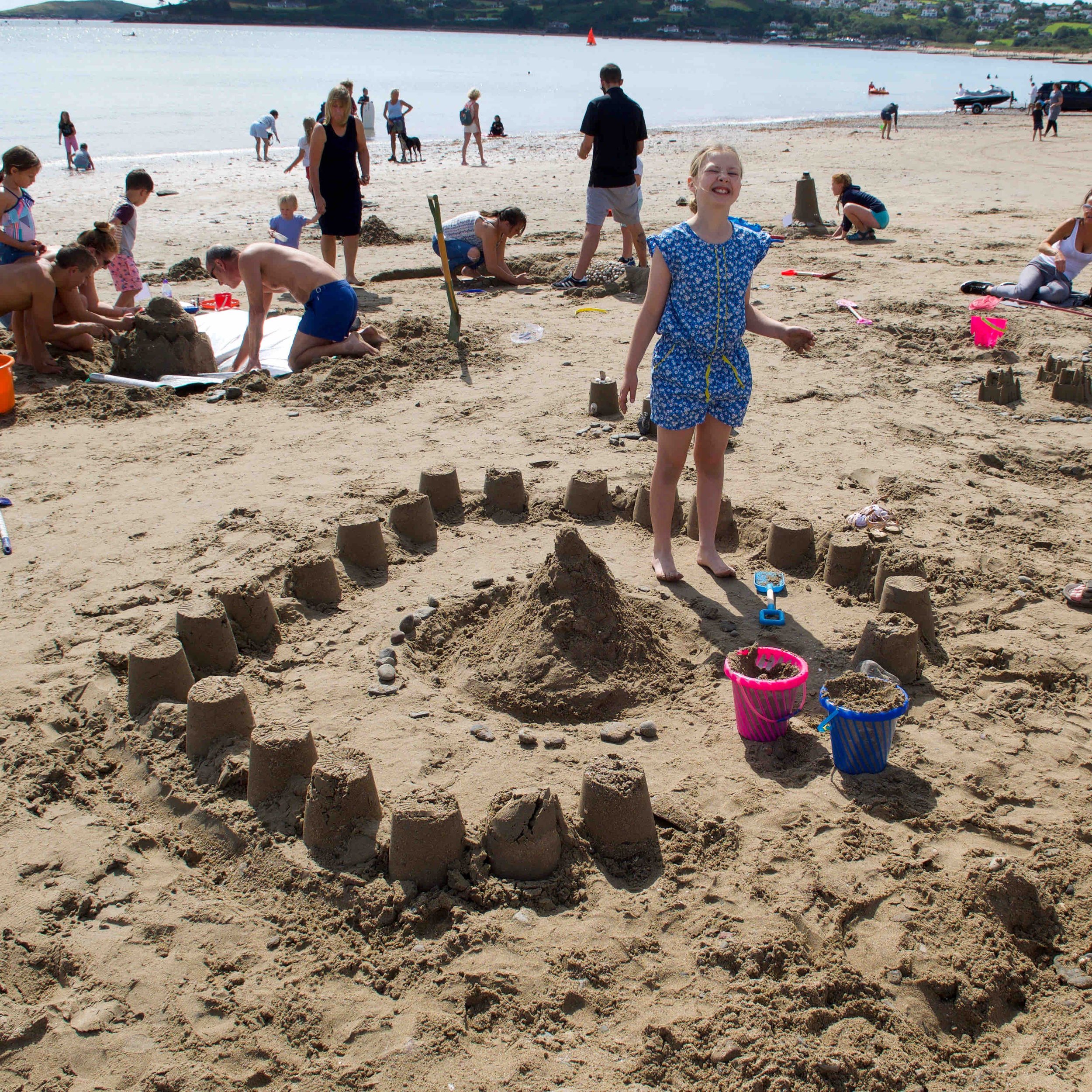 Children building sandcastles on a beach, with one child smiling next to a large sandcastle surrounded by pails. Other people are in the background, some sitting and building additional sand structures. The ocean is visible in the distance.