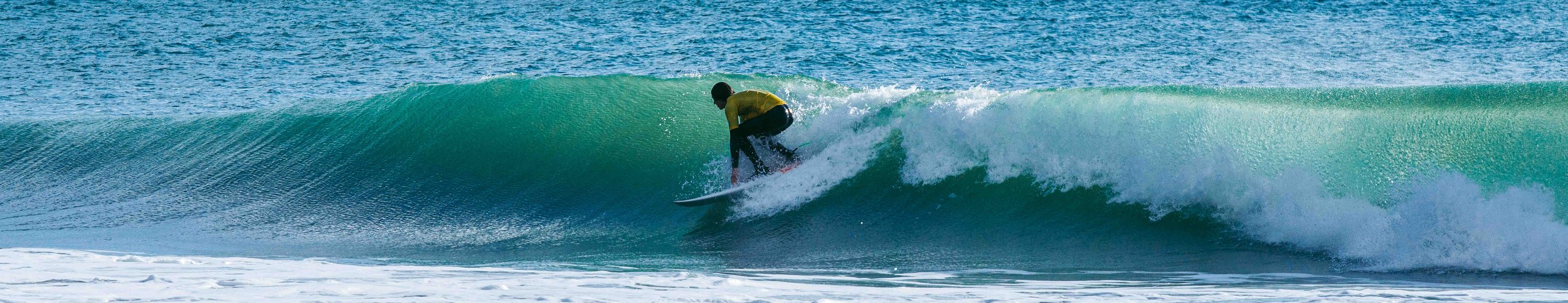 Surfer riding a wave in the ocean, wearing a yellow shirt and black wetsuit.