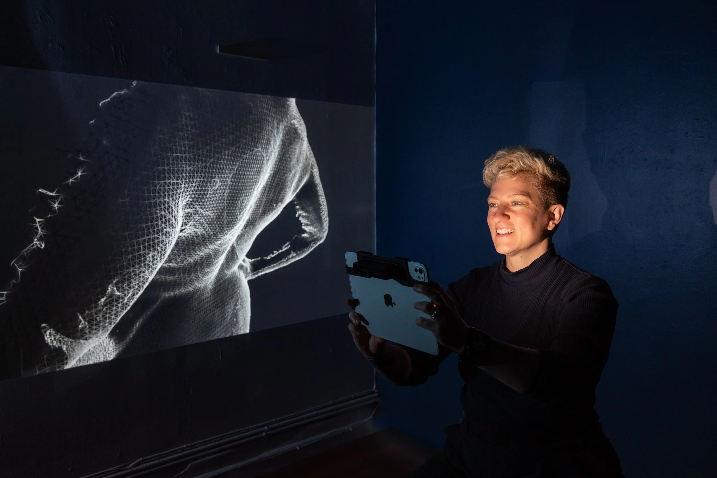 Lisa holding iPad with glow on her face in a dark room with a plus size back 3d scanned in mesh black and white as a 3d model