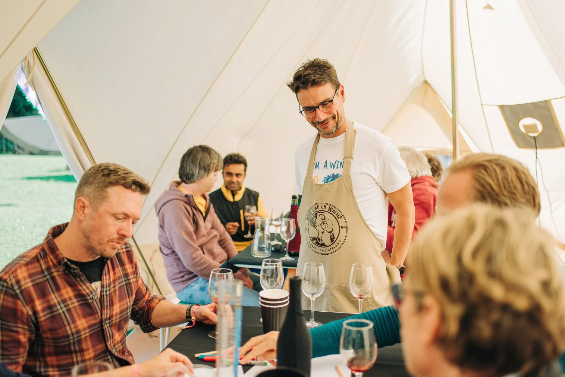 People sitting at a table inside a large tent at an outdoor event, with one person standing, wearing an apron and glasses, smiling.