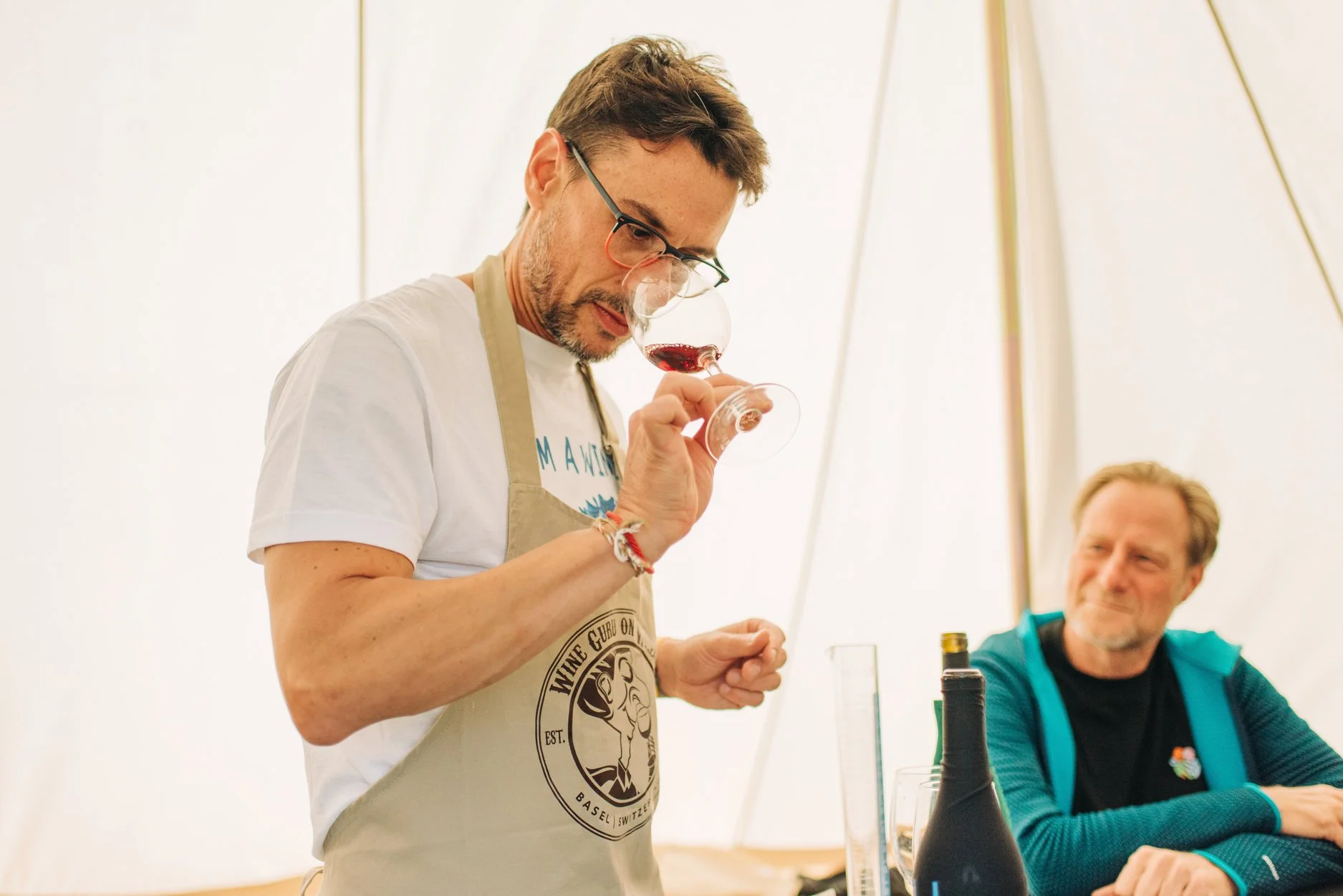 A man wearing glasses, a white shirt, and an apron tasting red wine in a wine tasting event while a man in a teal jacket observes and smiles in the background.