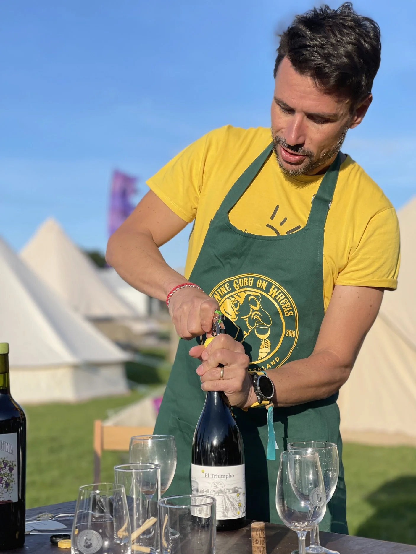 A man with dark hair, wearing a yellow t-shirt and green apron, opening a bottle of wine outdoors during daytime. There are wine glasses on the table, and in the background tents and a blue sky can be seen.
