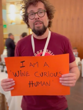 Person with curly hair and glasses holding an orange sign that says 'I am a wine curious human' in a conference or event setting.
