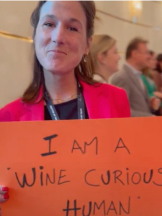 Person holding an orange sign that says, 'I AM A WINE CURIOUS HUMAN,' at a conference or event.