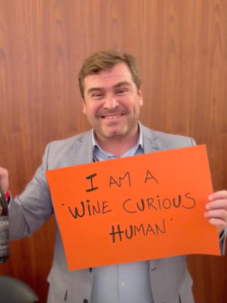 Man in a gray suit holding an orange sign that says 'I AM A WINE CURIOUS HUMAN' with a wooden wall background.
