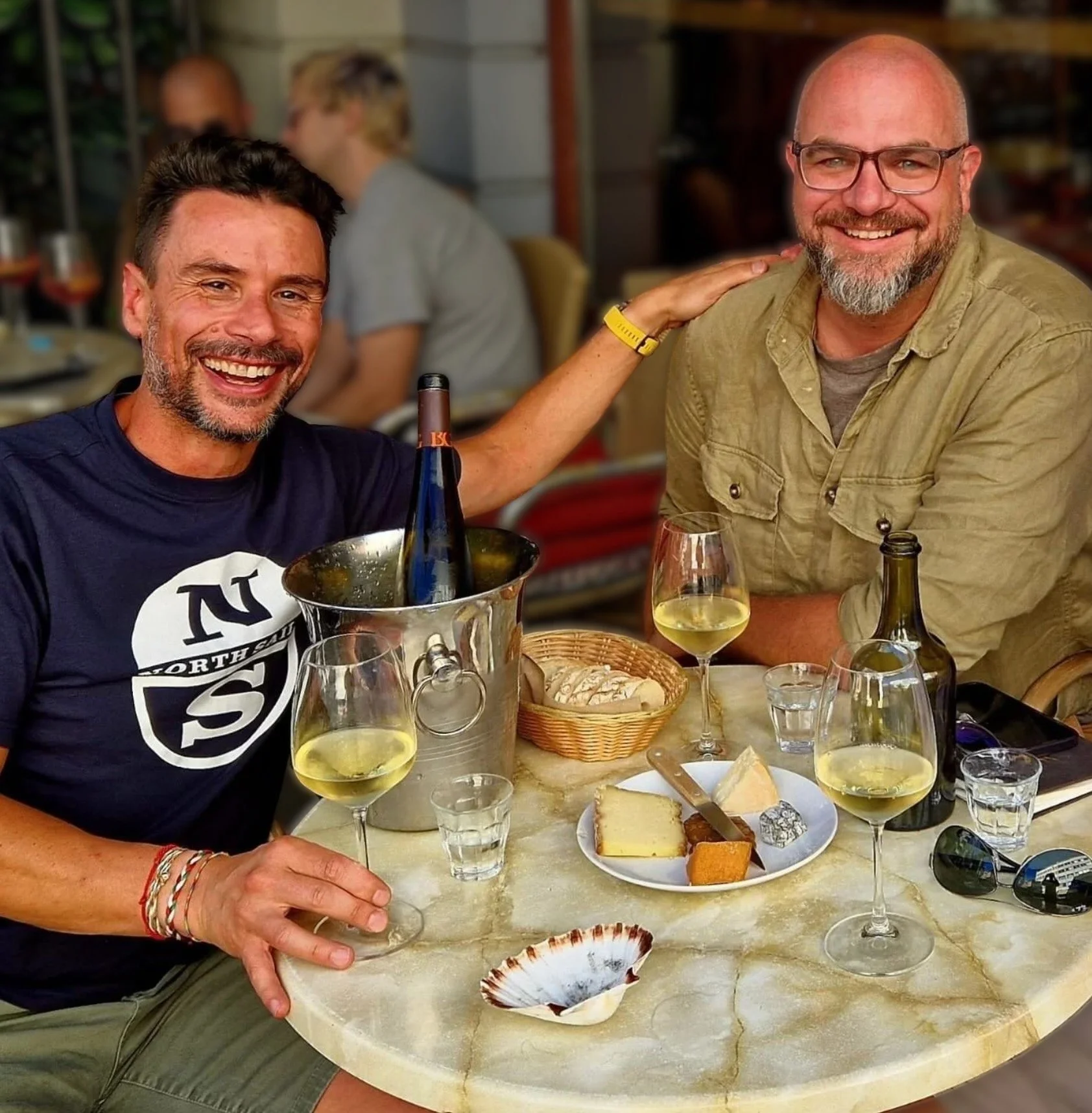Two smiling men at a dining table with wine glasses, cheese, bread, and snacks, enjoying a social gathering or wine pairing meal.