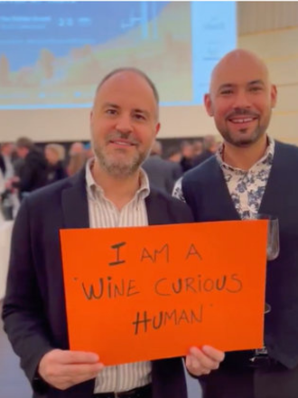 Two men in suits holding an orange sign that says, 'I AM A WINE CURIOUS HUMAN,' at a conference or event.