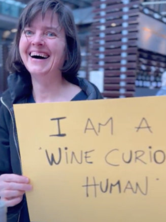 A woman holding a yellow sign that reads 'I AM A WINE CURIOUS HUMAN' with a happy expression