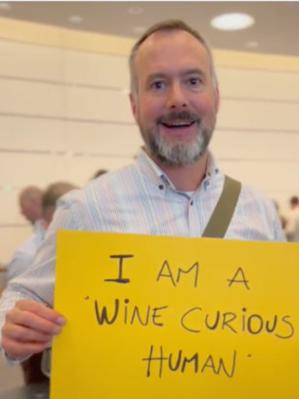 Man smiling at an event holding a yellow sign that reads 'I AM A WINE CURIOUS HUMAN'.