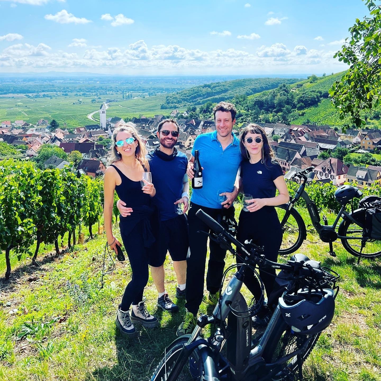Group of five friends smiling with bicycles in a vineyard overlooking a small European village and rolling hills on a sunny day.