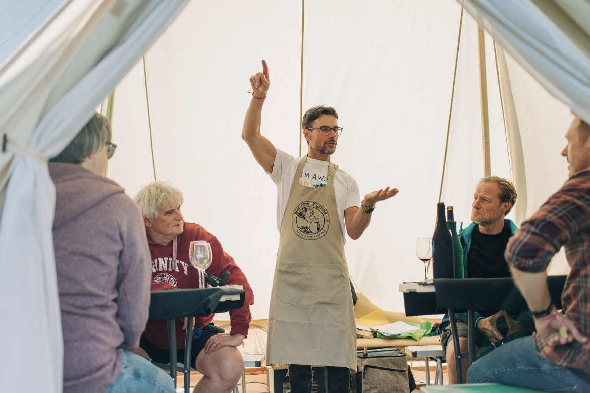 A man wearing glasses and an apron is giving a presentation or speech inside a tent to four seated adults, during a wine tasting or demonstration event.