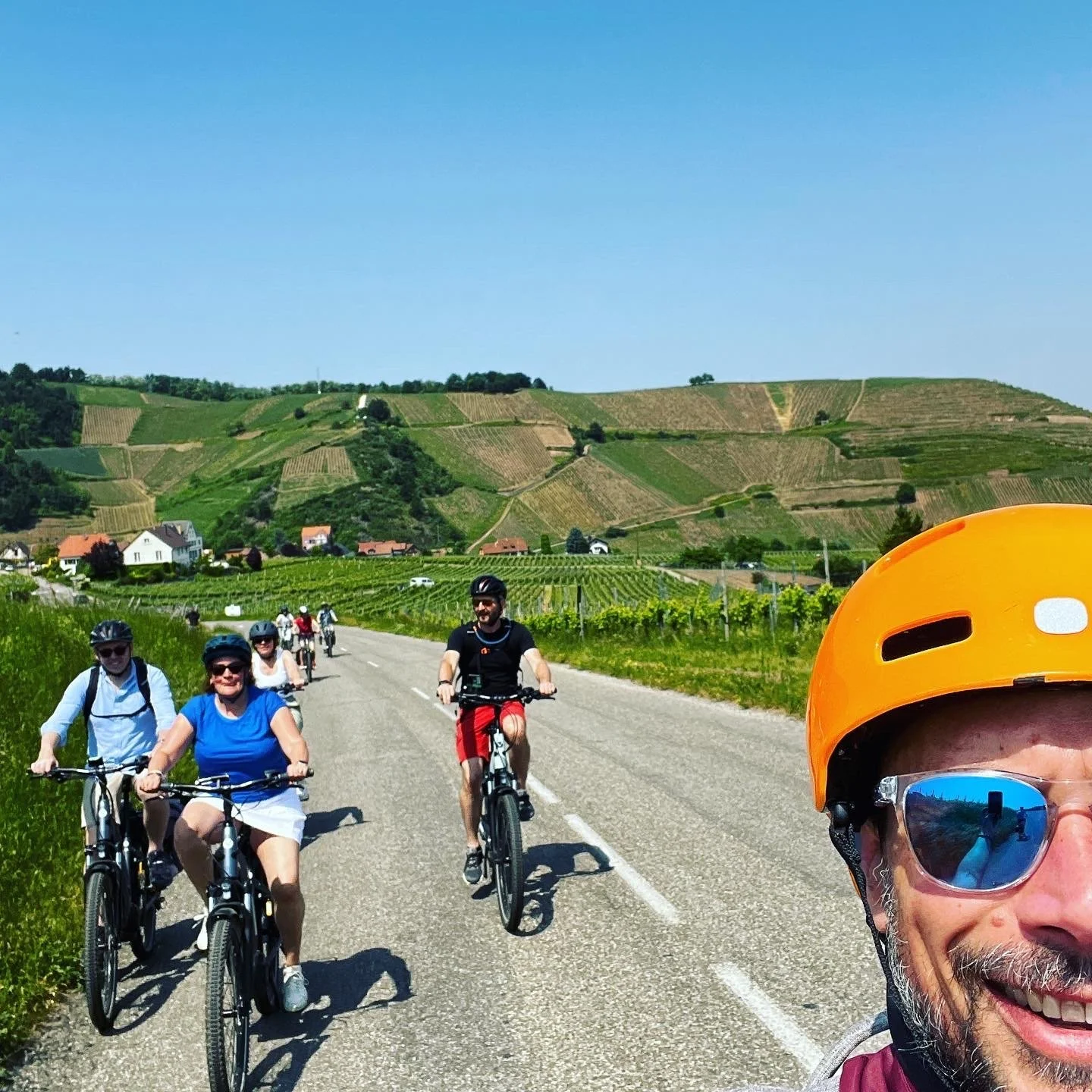 A group of people cycling on a rural road surrounded by vineyards and green hills under a clear blue sky, with one person in the foreground wearing an orange helmet and sunglasses.