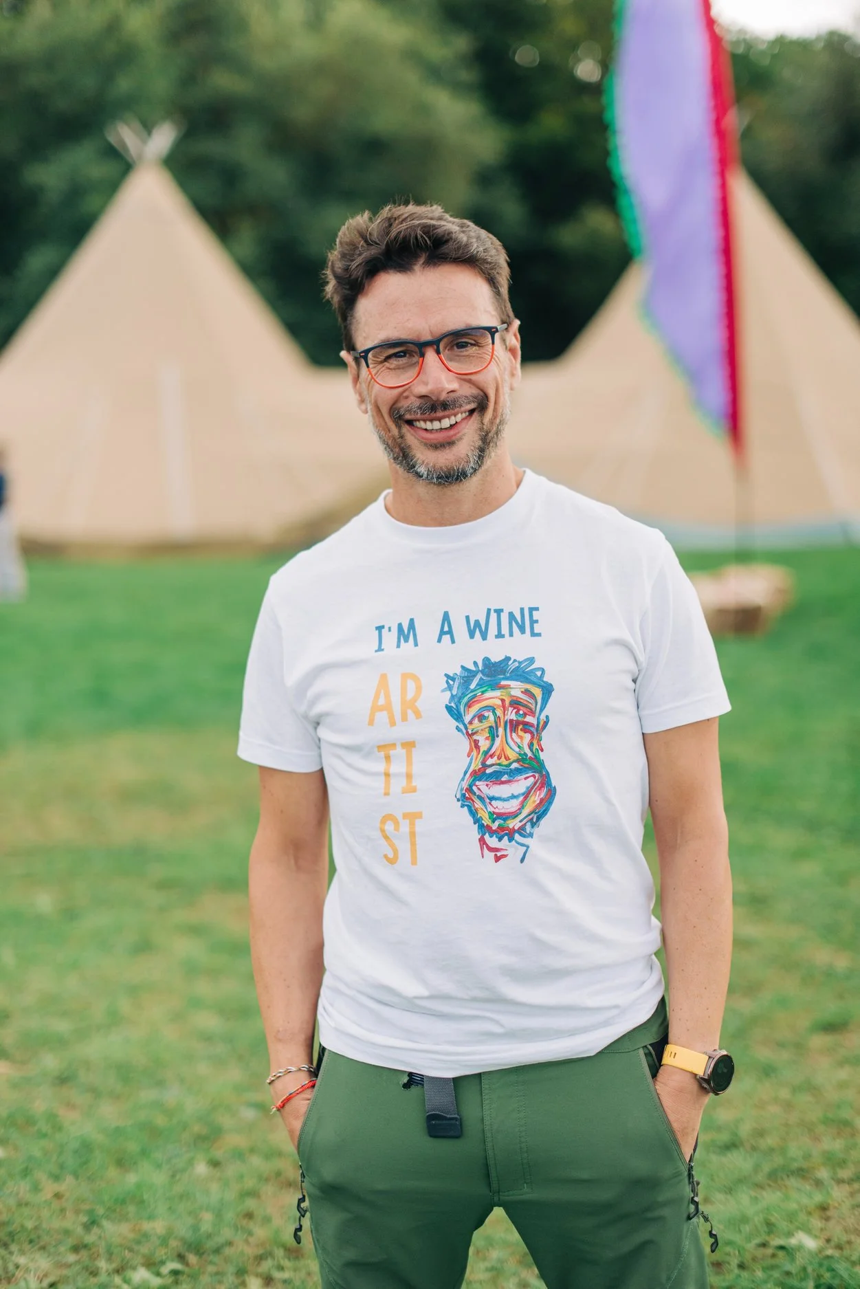 A smiling man with glasses, wearing a white T-shirt with colorful text and a face illustration, standing outdoors in front of teepees and a colorful flag.