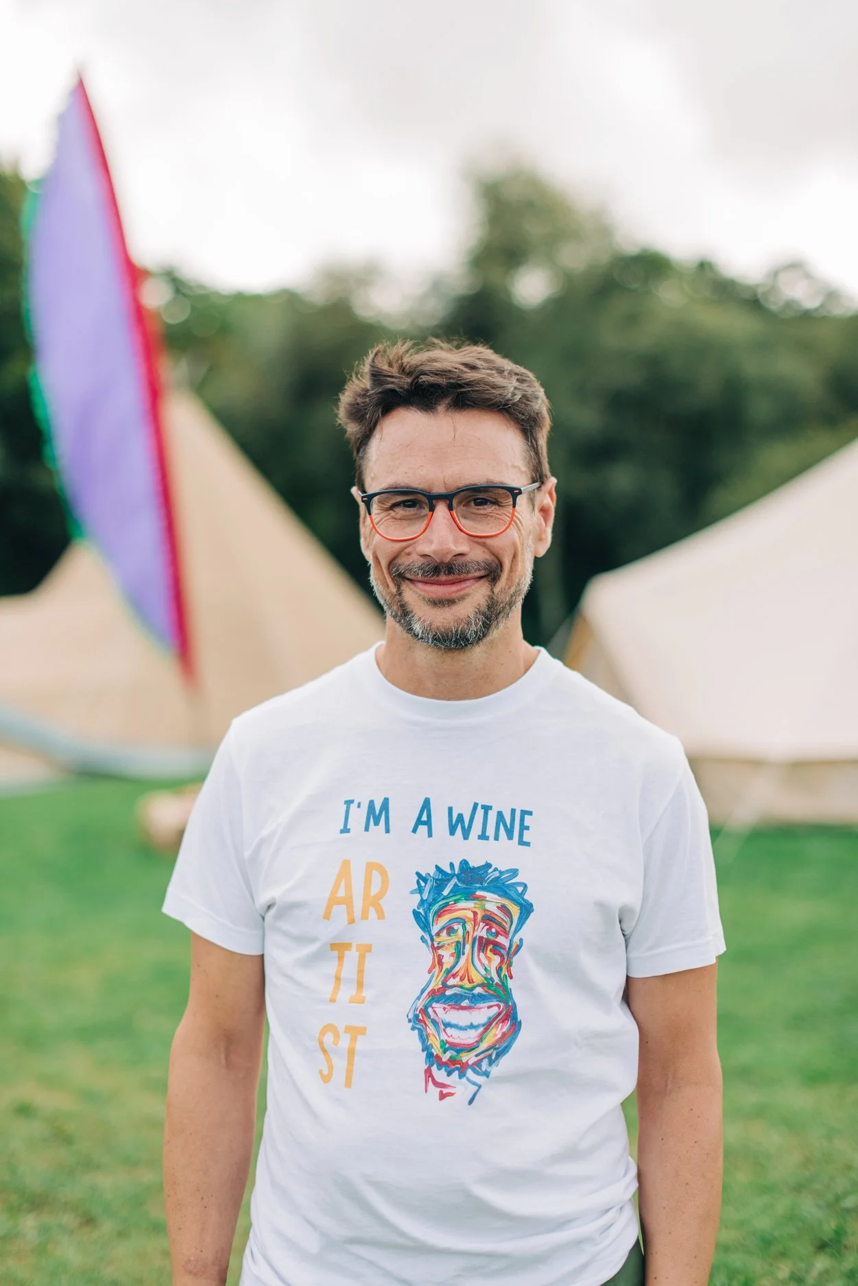 A smiling man with glasses standing outdoors in front of tents and a rainbow-colored umbrella during daytime.