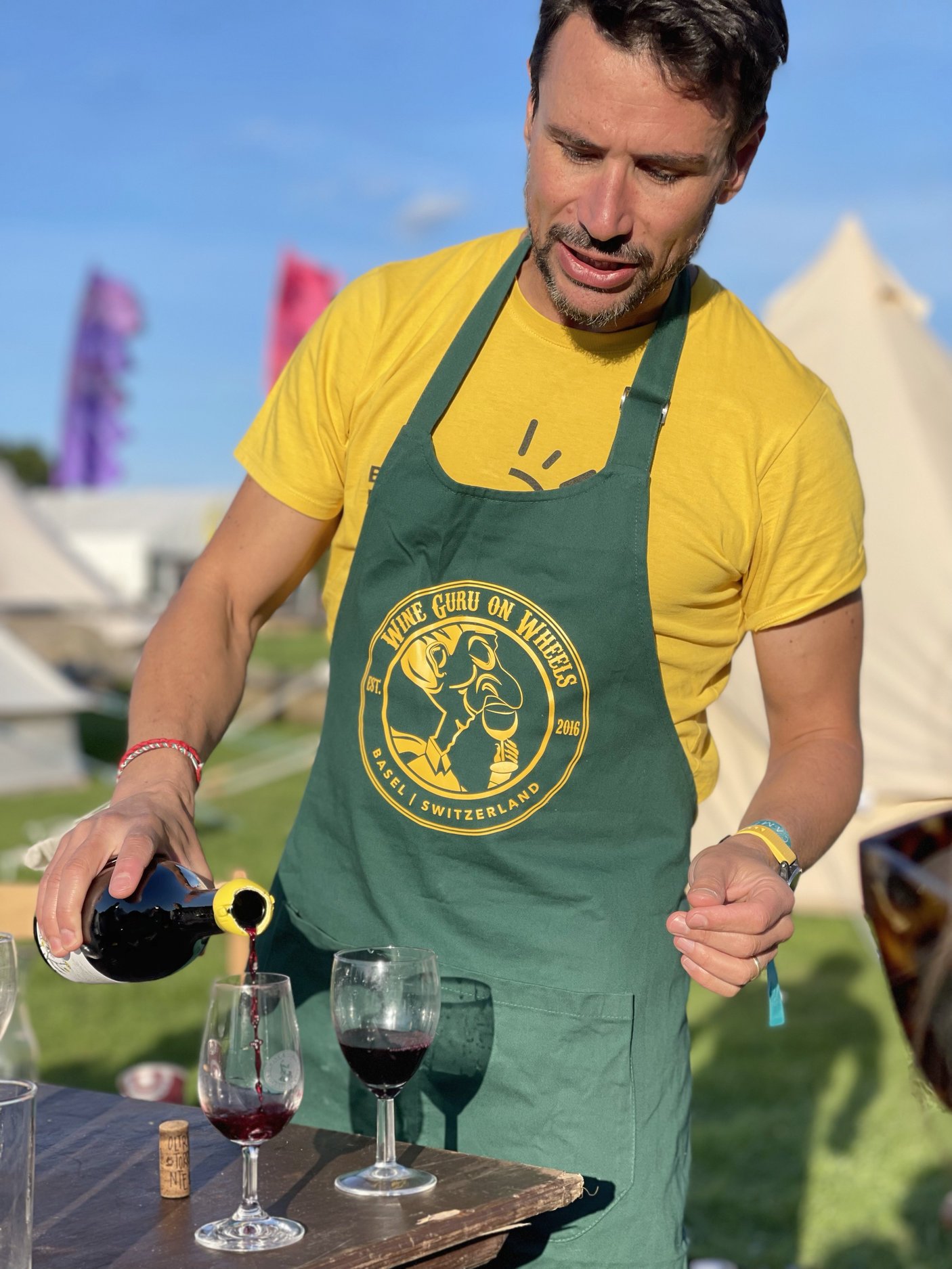 A man in a yellow t-shirt and green apron pours red wine into glasses on a wooden table outdoors during daytime.