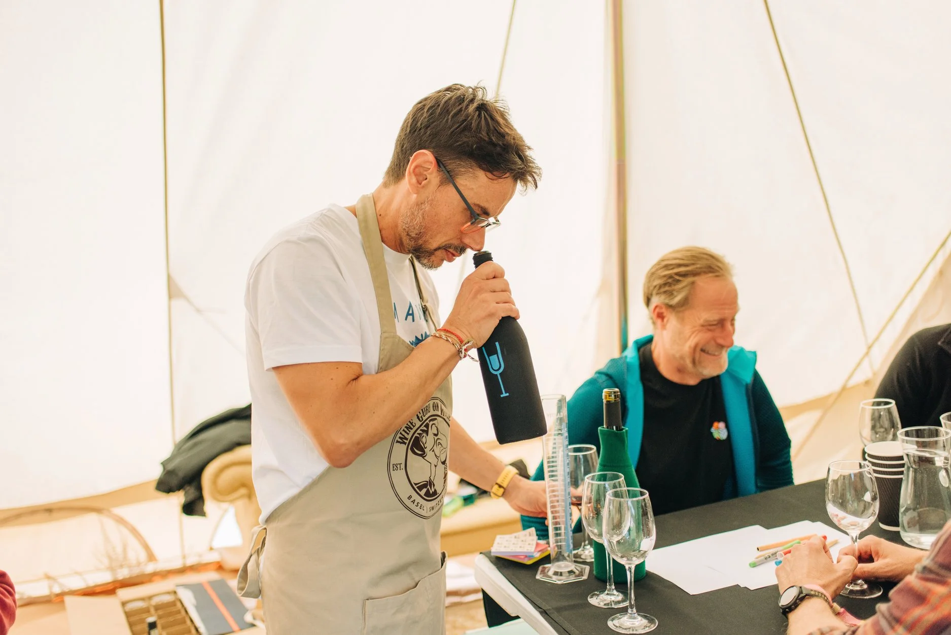 Man with glasses tasting wine at a wine tasting event, wearing a beige apron and white t-shirt, smiling man in blue jacket and black shirt, table with glasses and wine bottles, in a tent.