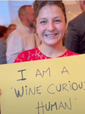 A smiling young woman holding a yellow sign that reads "I am a 'wine curious' human" at a gathering or event.
