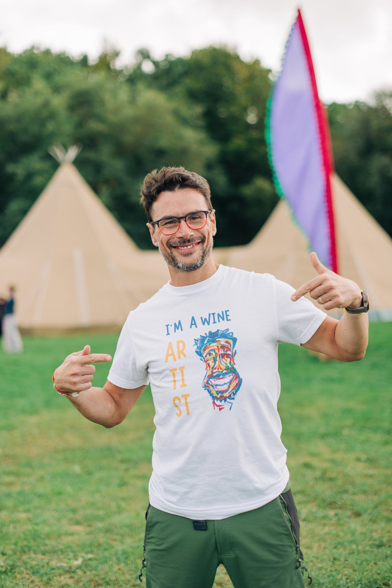 A man with glasses smiling and pointing to his graphic T-shirt that reads, 'I'M A WINE ARTIST,' with a colorful abstract face illustration, standing outdoors in front of tents with trees in the background.