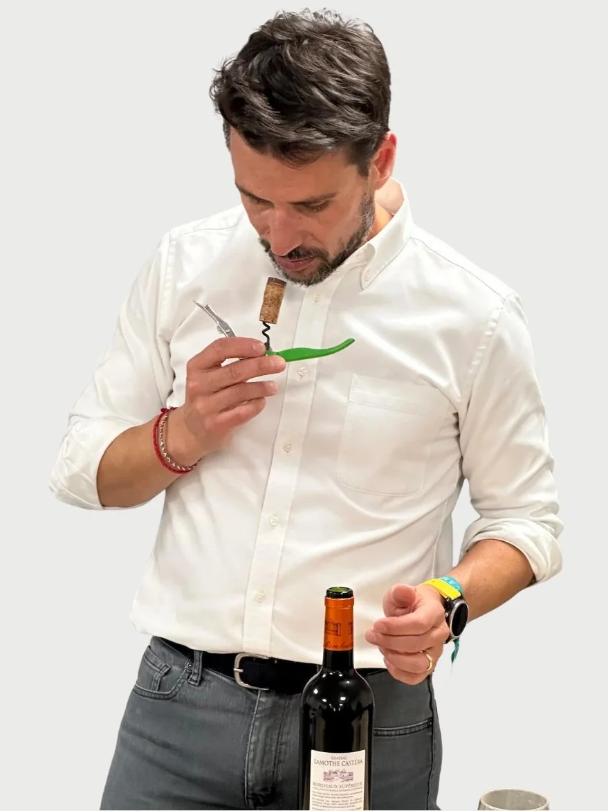 A man wearing a white shirt and watch holding a corkscrew and preparing to open a bottle of wine, smelling the wine for faults.