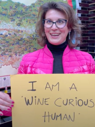 A smiling woman wearing glasses and a pink jacket holding a sign that says, 'I AM A WINE CURIOUS HUMAN.'