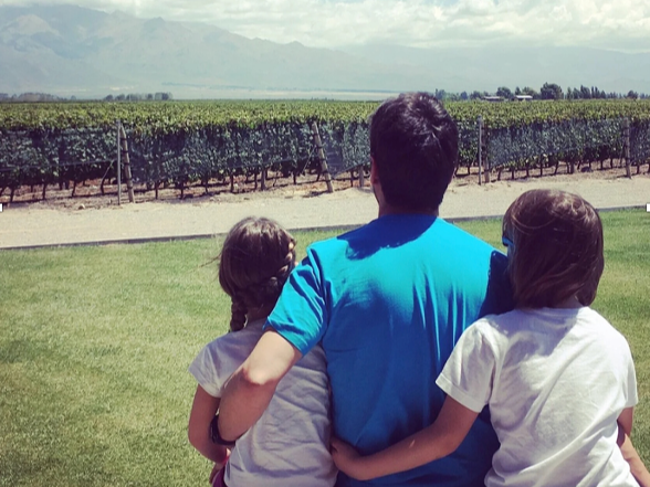 Two children sitting on the grass with their dad and looking at a vineyard with green vines and a cloudy sky.