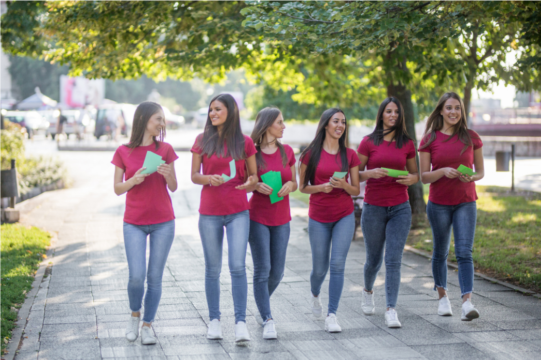 Group of six women in red shirts and jeans walking on a paved path, carrying green papers, under trees in a park-like setting.