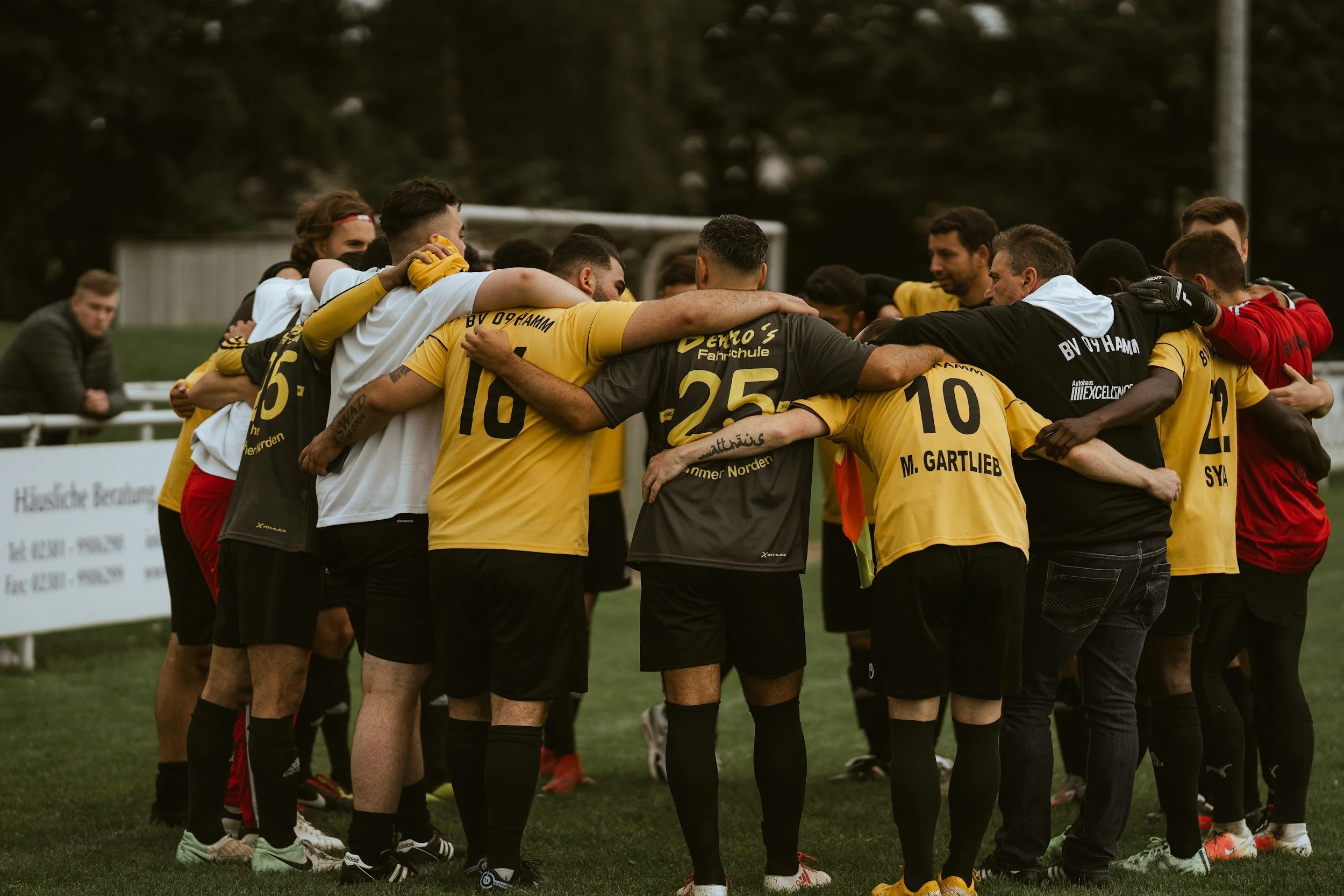 Soccer team huddled together during a game or practice, with players wearing yellow and black jerseys, and some wearing gloves, on a grassy field.
