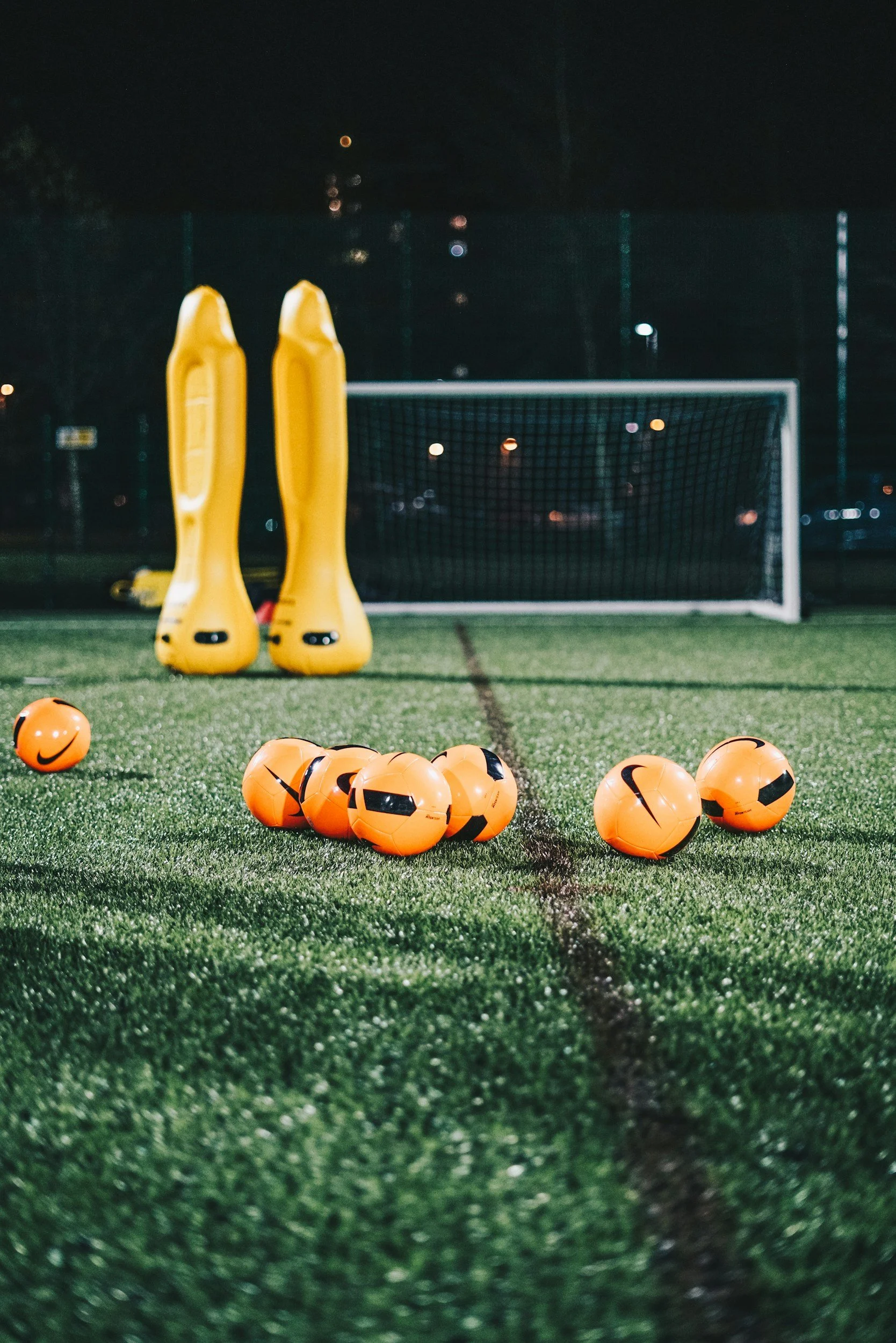 Soccer field at night with orange soccer balls, yellow training dummies, and a goal in the background.