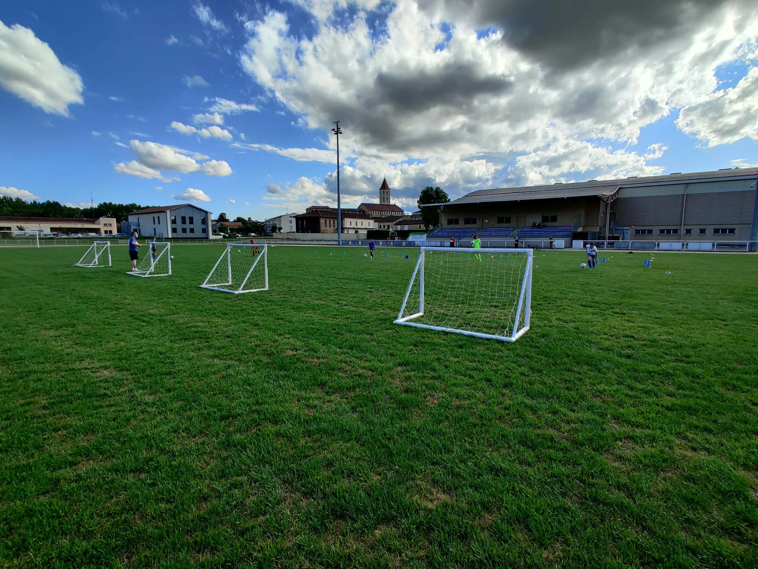 Soccer practice on a field with small goal nets and players standing near cones, under a partly cloudy sky with a building and trees in the background.