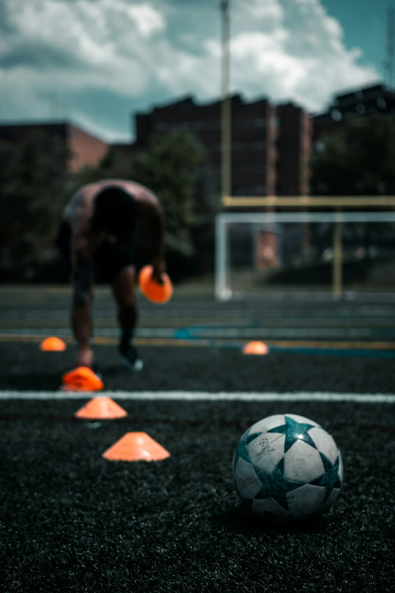 A soccer ball in the foreground on a soccer field with a person practicing drills in the background, marked by orange cones, under a cloudy sky.
