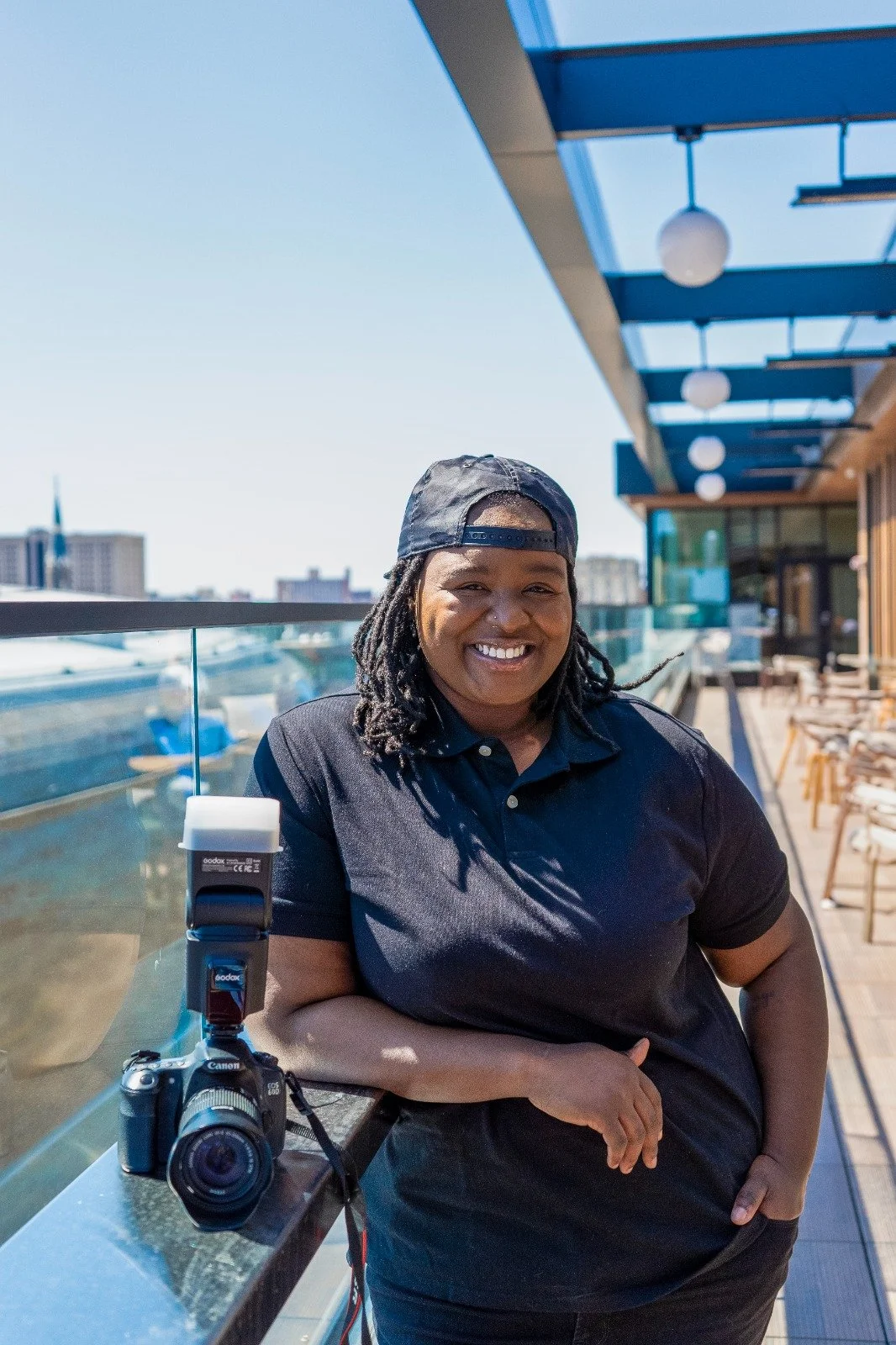 A woman with dreadlocks wearing a black cap backward and a black shirt, smiling on a rooftop terrace with city buildings in the background. A camera with a flash is on the table next to her.
