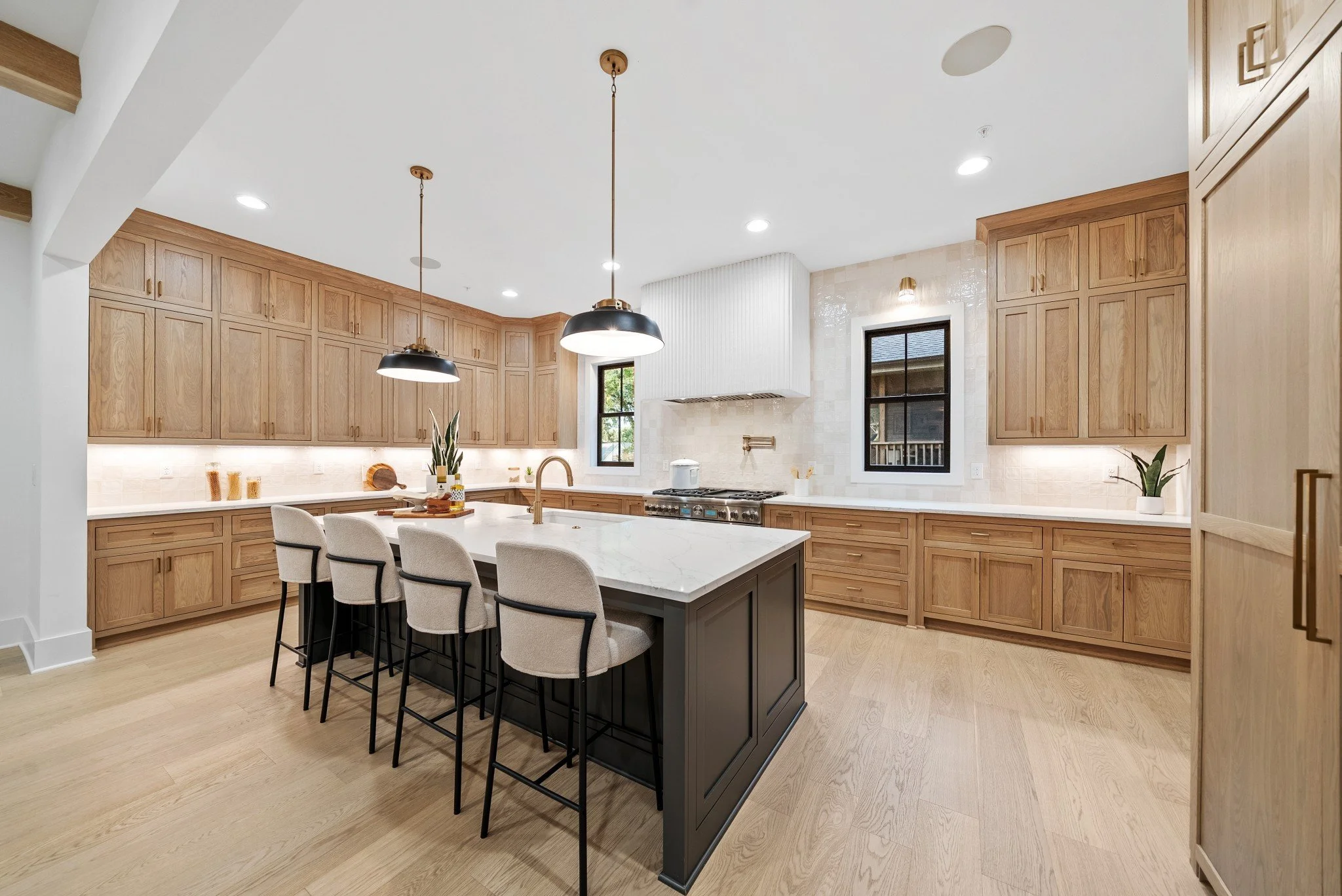 Modern kitchen with light wood cabinets, a large island with a dark base, white marble countertop, pendant lights, and two black-framed windows.
