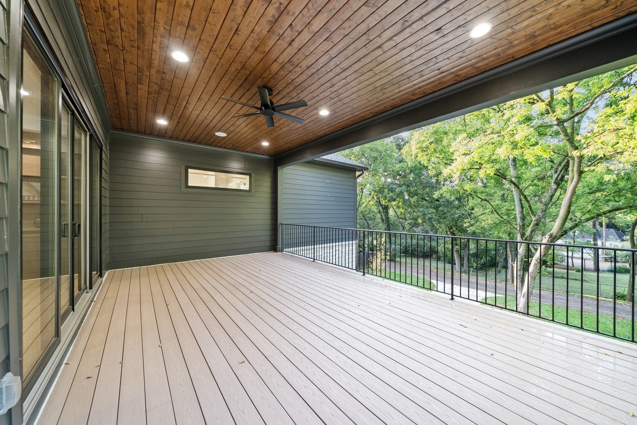 Newly built outdoor deck with wood planks, ceiling fan, and black metal railing, overlooking a lush backyard with trees.