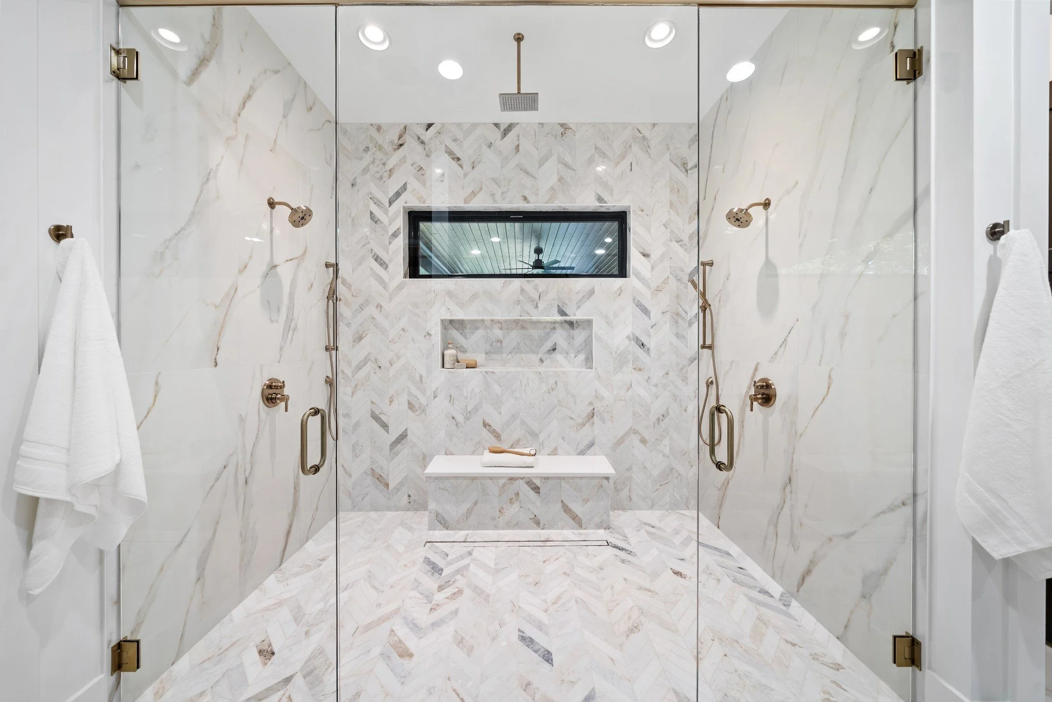 A modern glass-enclosed shower with marble walls and floor, featuring two shower heads, a built-in niche with toiletries, and a rectangular window.