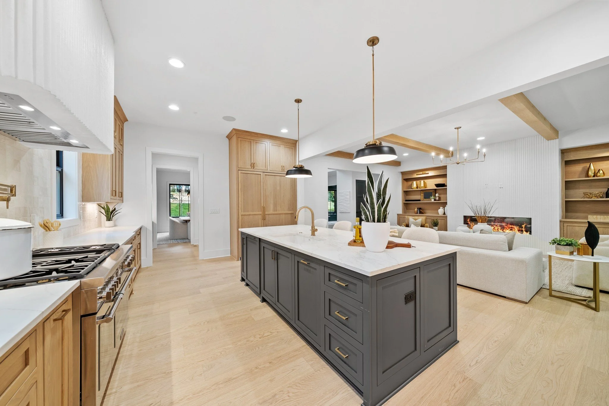 Modern open-concept kitchen and living room with light wood flooring, white walls, a large kitchen island with a dark base and white marble countertop, pendant lighting, and a cozy fireplace area with built-in shelving and seating.