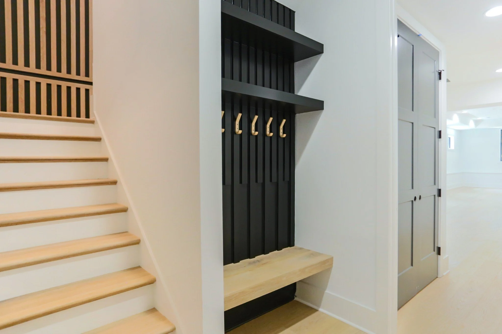 Interior view of a modern home with wooden staircase, black wall hooks, a bench, and gray doors.