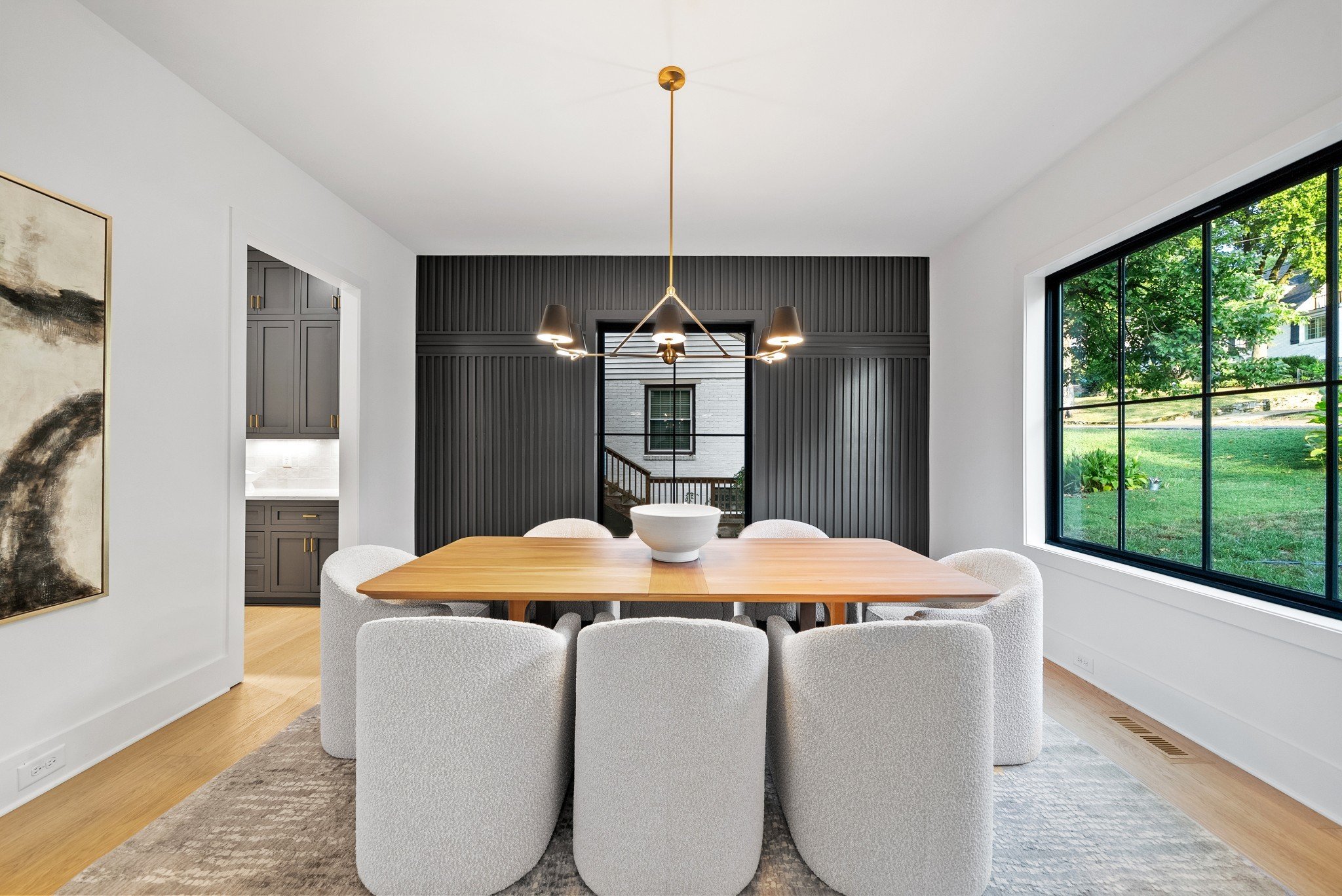 Modern dining room with a wooden table, white upholstered chairs, a black accent wall with a door, large window showing green outdoor scenery, with a gold chandelier overhead and a large abstract painting on the white wall.