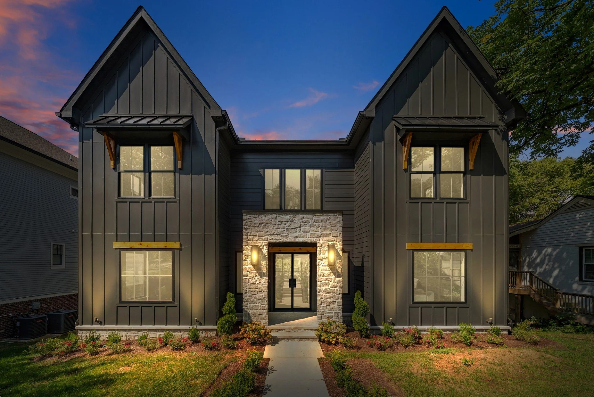 Modern two-story house with dark exterior, stone accents at entrance, surrounded by a well-maintained lawn and garden, during sunset.