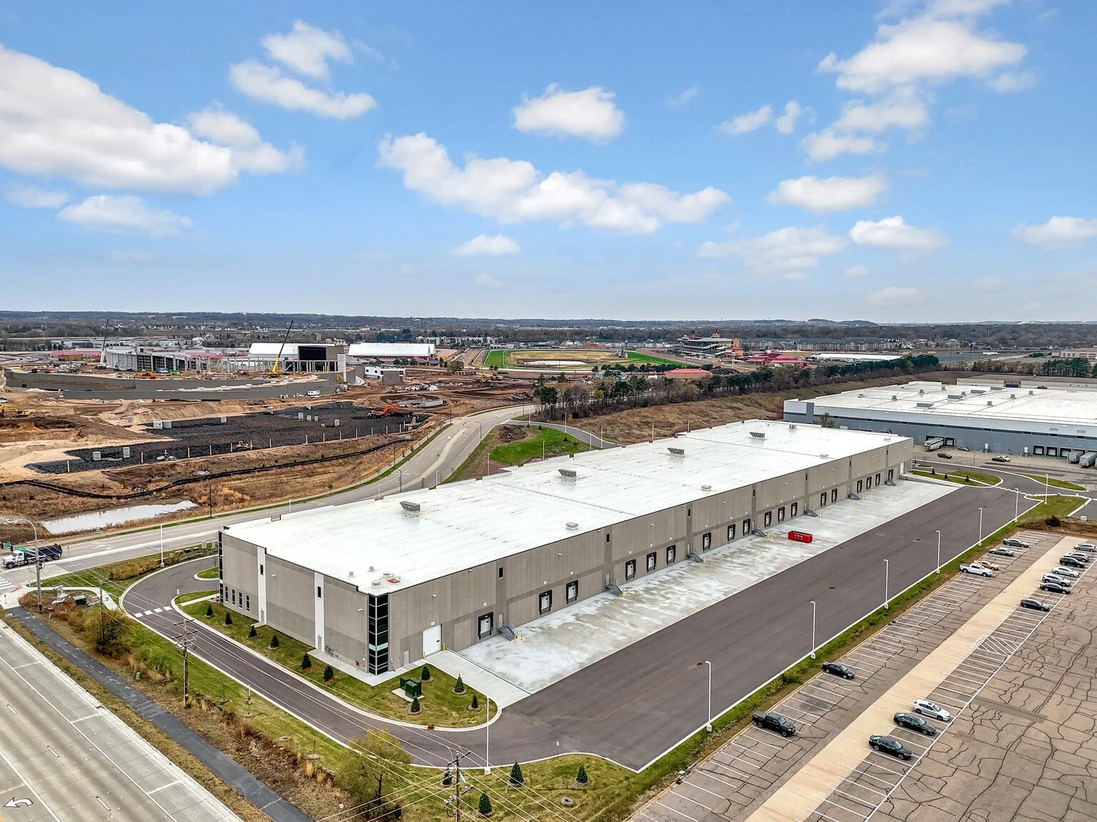 Large industrial warehouse building with white roof, adjacent parking lot with cars, surrounded by paved roads and green lawns. Construction site visible in background.