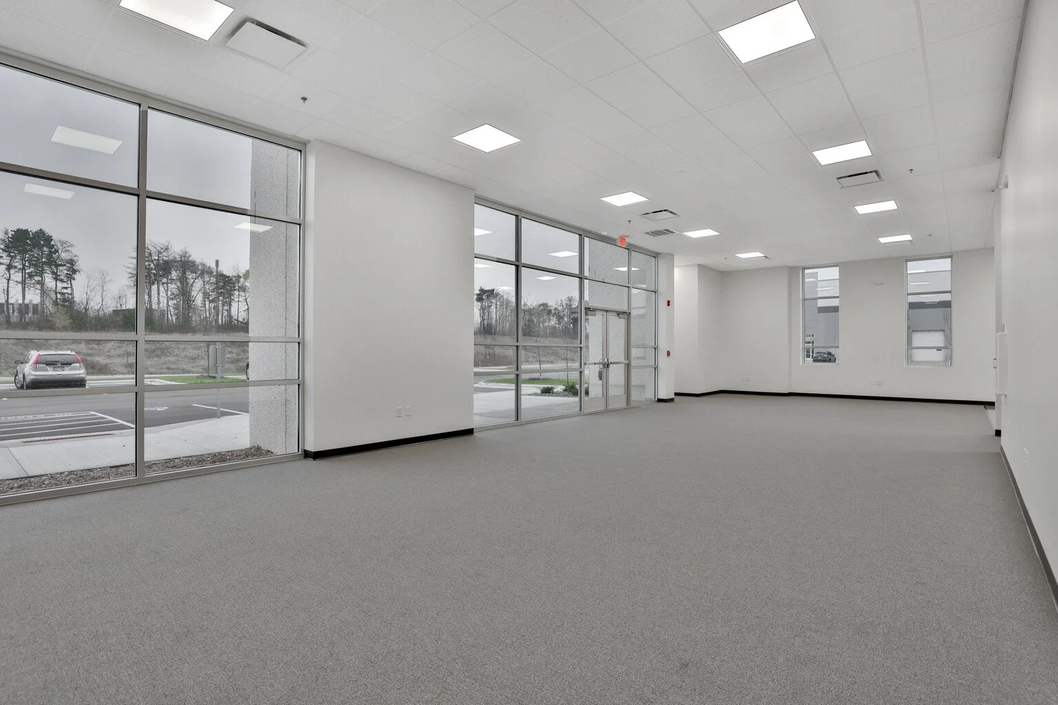 Commercial space with large windows, white walls, ceiling lights, and gray carpeted floor.