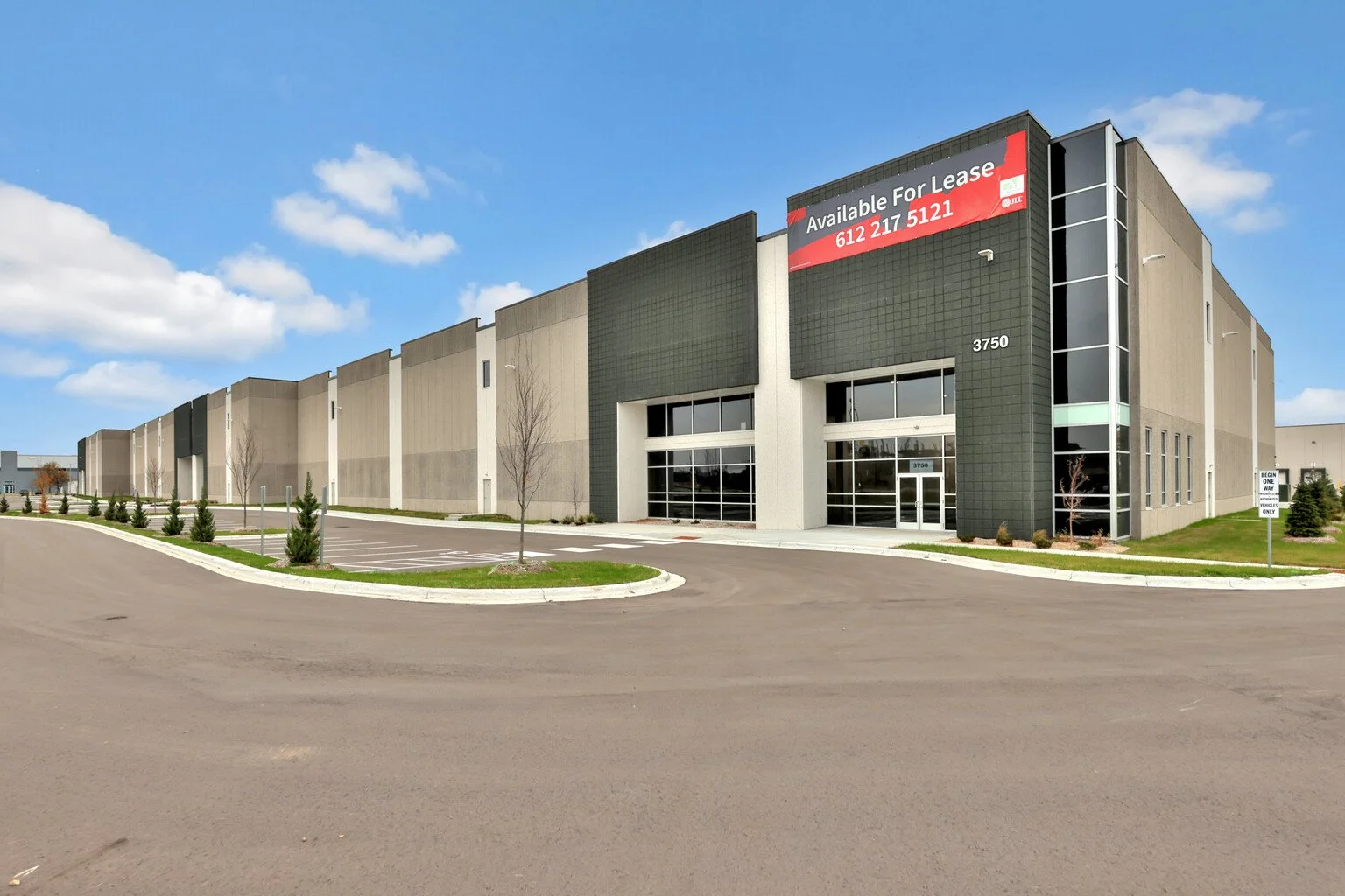 Modern commercial building with large glass entrance and an electronic sign reading "Available for Lease" with a phone number. Parking lot with a few small trees and a clear blue sky.
