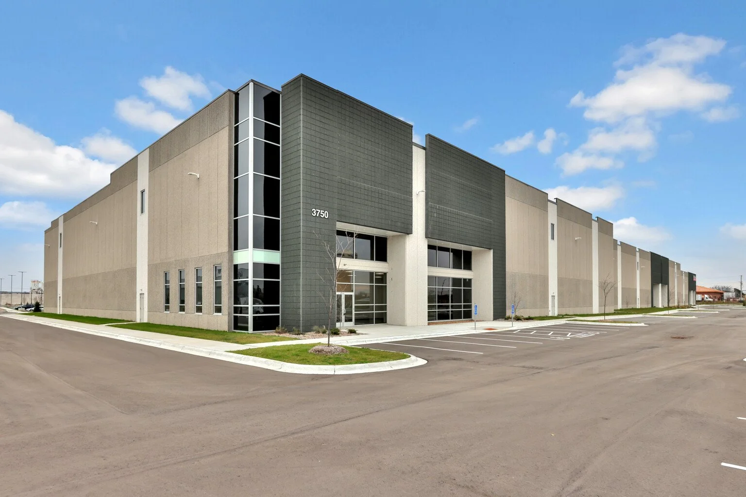 Empty parking lot in front of a modern commercial building with gray and beige exterior walls and large glass entrance, under a partly cloudy sky.