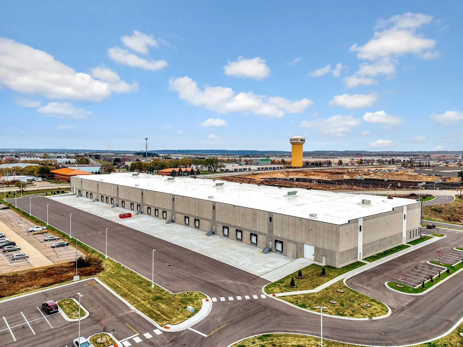 An aerial view of a large industrial building with a white roof, surrounded by parking lots, roads, and a partly cloudy sky.