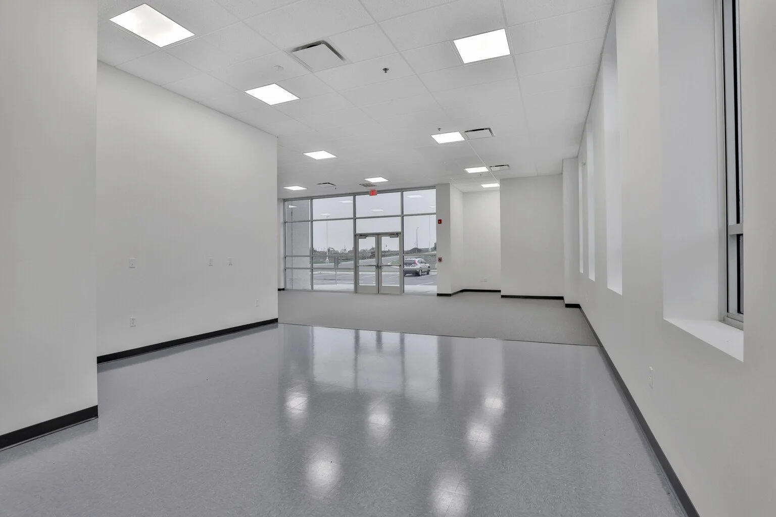 Empty commercial space with gray flooring, white walls, large windows, and an entrance door, with a parking lot visible outside.