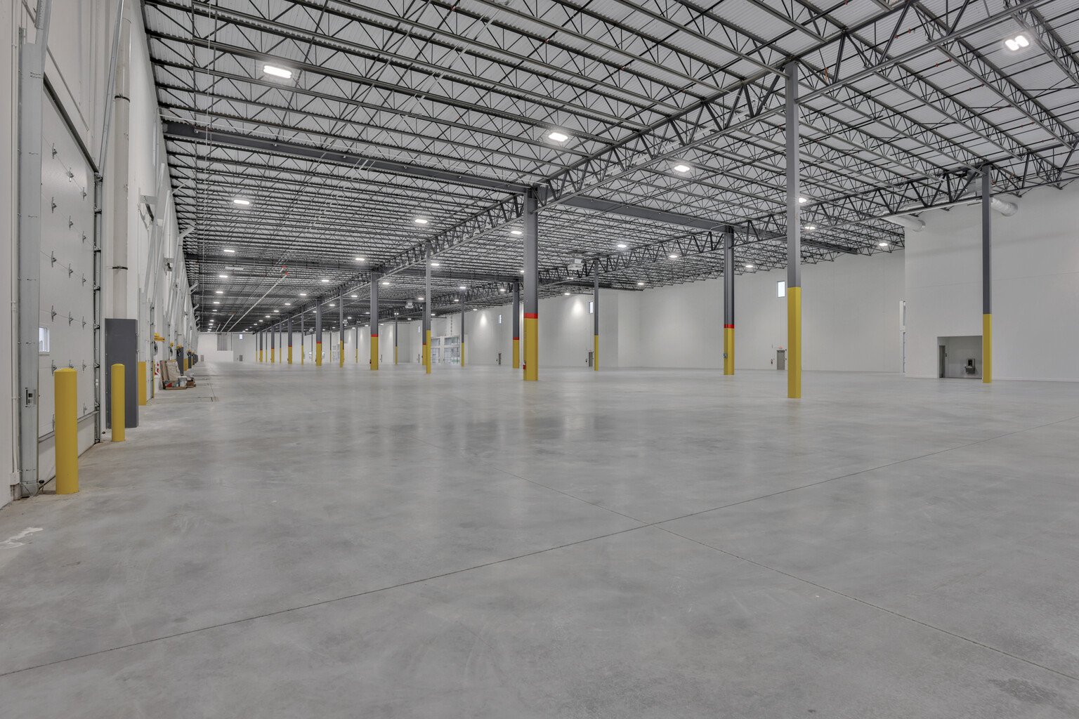 Empty warehouse with high ceiling, metal support beams, concrete floor, and roll-up doors on the left wall.